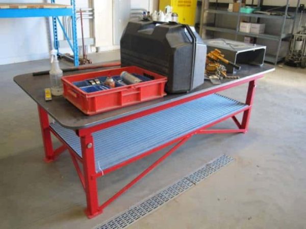 Red heavy duty welding table with a steel top and materials on the surface in a warehouse setting.