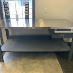 Grey workbench with a stainless steel top, drawer, and lower shelf in an office setting with a key taped to the front of the drawer.